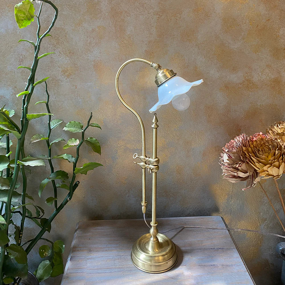 Vintage-style brass lamp with white glass shade on a wooden surface, next to a plant and flowers.