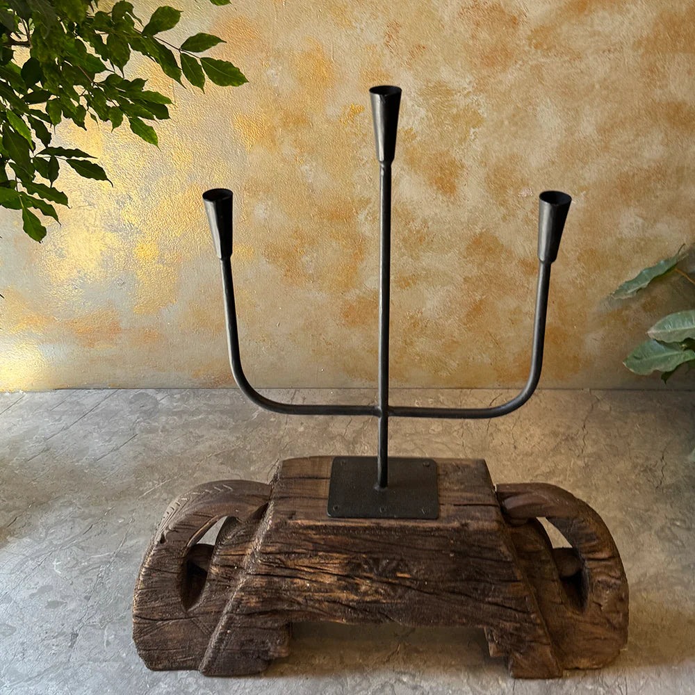 Decorative candle holder with three candlesticks on a wooden base against a textured wall.