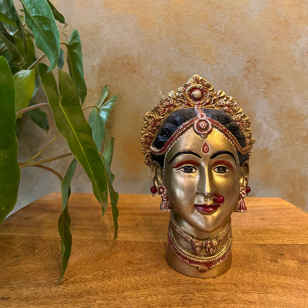Decorative statue of a face with jewelry on a wooden surface with a plant in the background