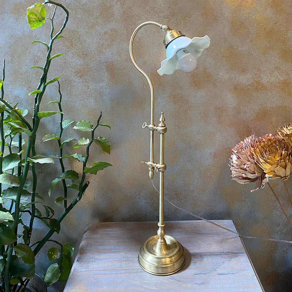 Vintage-style brass lamp with a white floral shade on a wooden surface, with plants and flowers in the background.