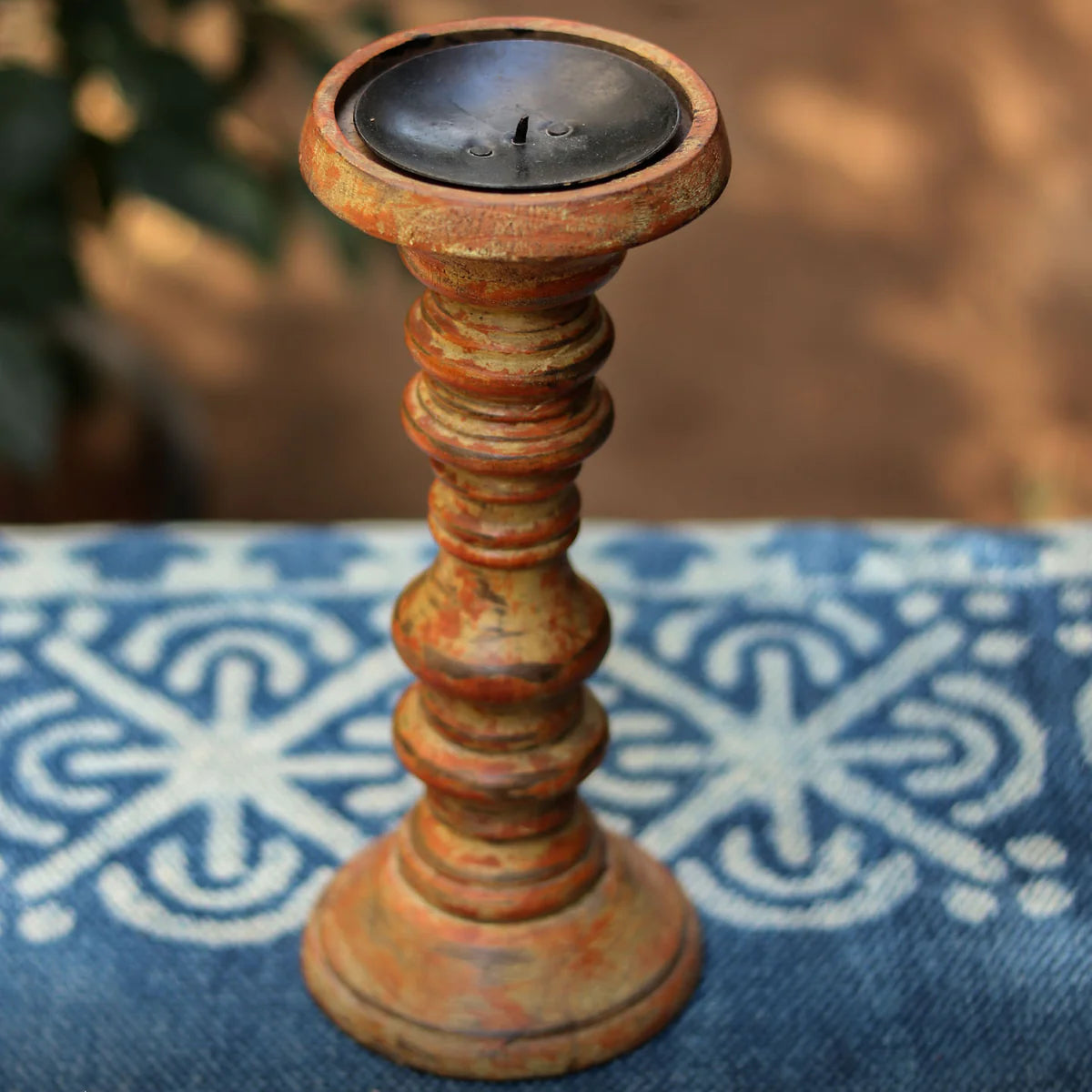 Rustic candle holder with a candle on a blue patterned surface, blurred greenery in the background.