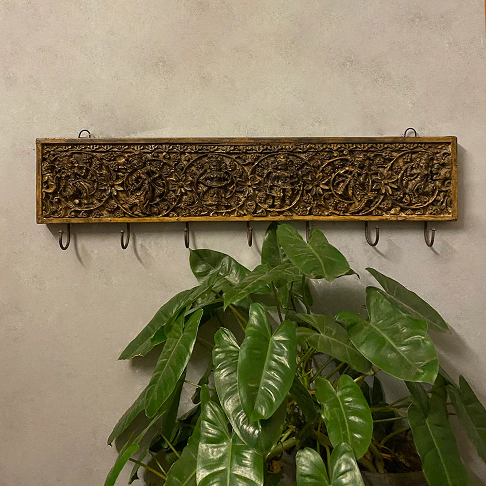 Decorative wooden wall hook with intricate carvings and a plant below on a light gray wall.