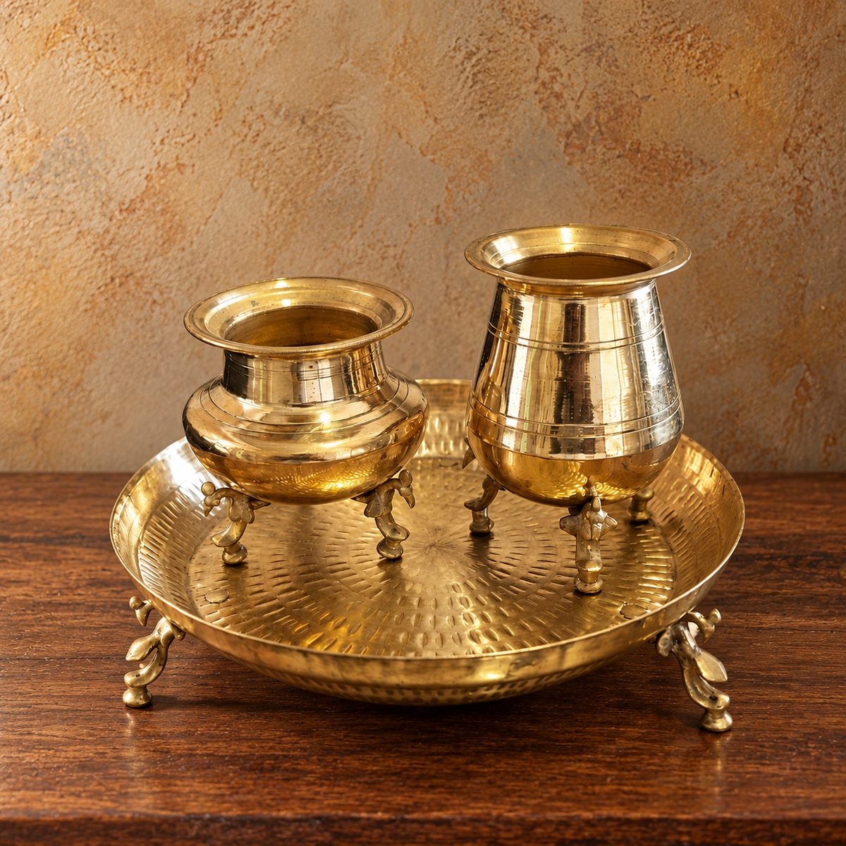 A brass three-piece set including a deep bowl, a small cup, and a hammered tray, displayed on a dark wooden table against a textured beige background.