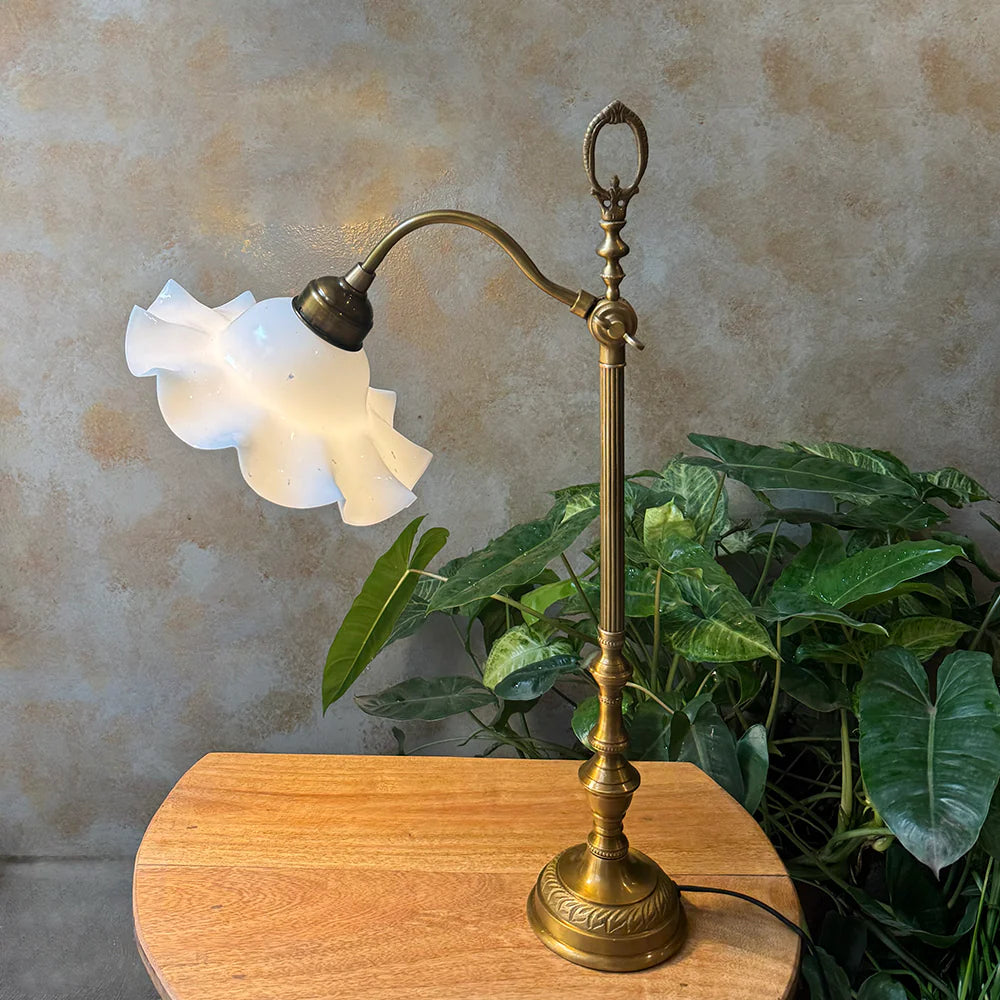 Vintage-style brass lamp with white glass shade on a wooden surface with plants in the background.