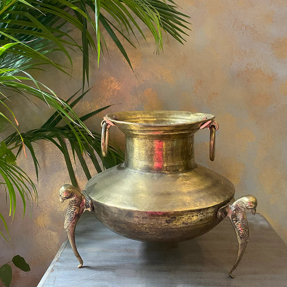 An aged brass pot, featuring three peacock-shaped feet and side rings, is filled with a bouquet of dried pink flowers.